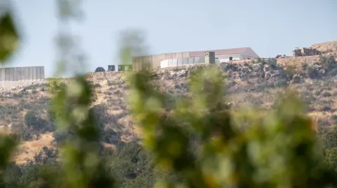 Phil Caller A distant view of a building and trucks on the edge of a mountain