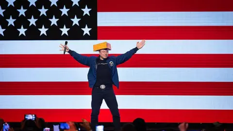 Getty Images Musk stands in front of a huge US flag and wears a cheese-shaped hat