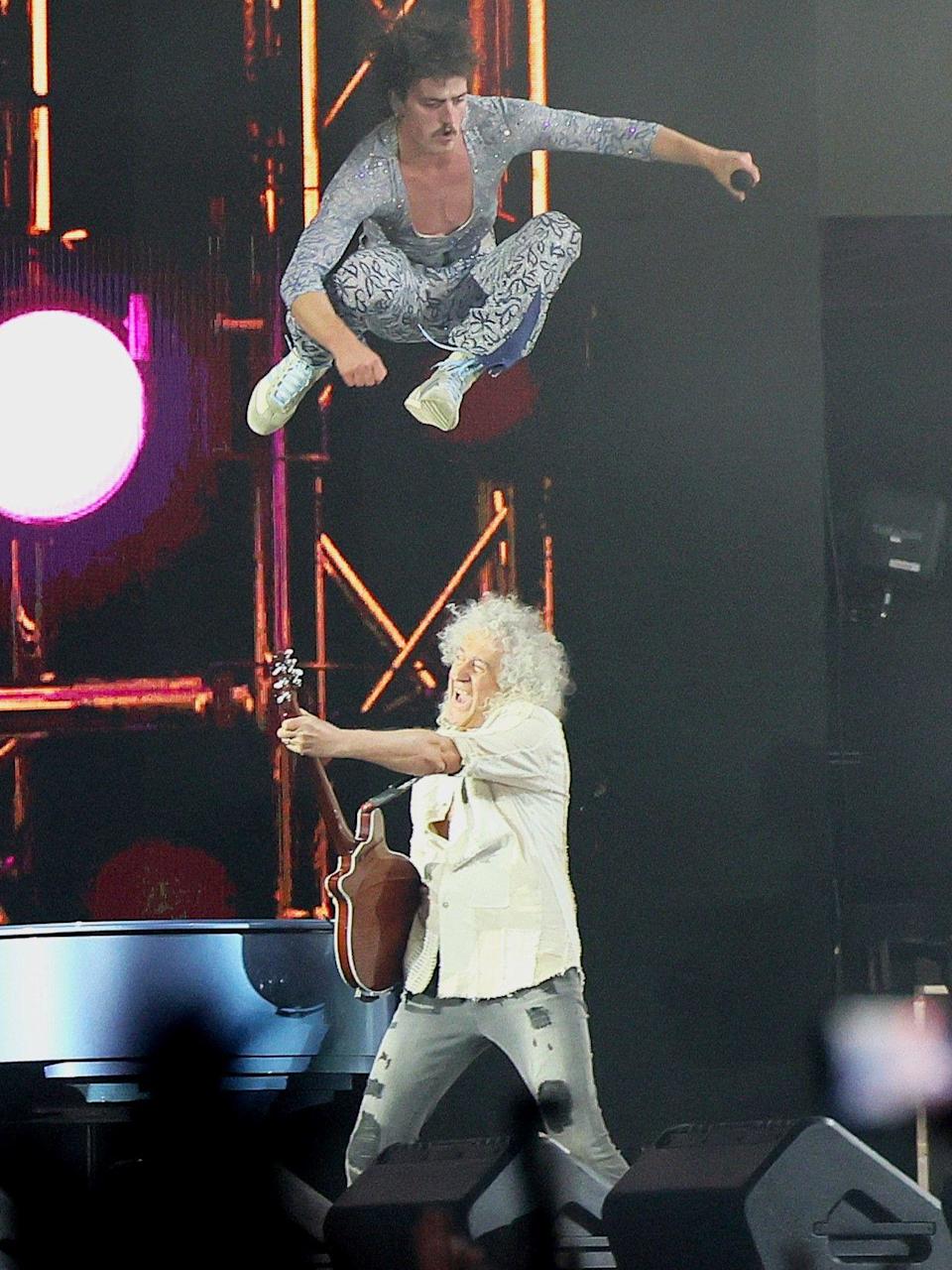 Benson Boon, wearing a blue leotard, flips over Brian May while he plays the guitar on stage.