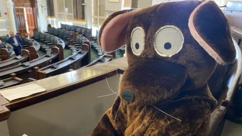 LDRS A person in a rat costume sits above the council floor at the meeting on Tuesday.