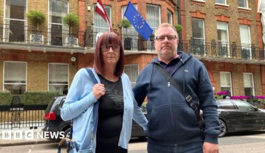 A man and woman standing on a street in London. The woman has shoulder-length red hair and is wearing a blue cardigan. The man is in a navy zip-up hoodie and they both have their arm around each other. There is an EU flag behind them and black parked cars.