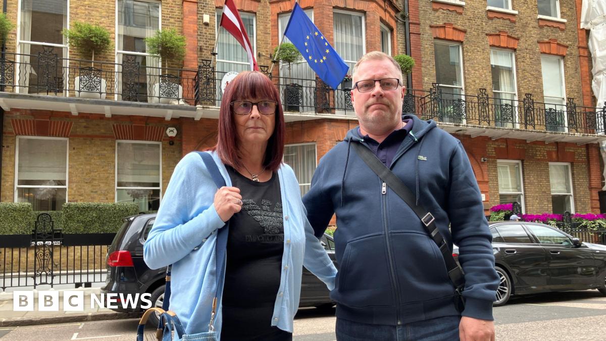A man and woman standing on a street in London. The woman has shoulder-length red hair and is wearing a blue cardigan. The man is in a navy zip-up hoodie and they both have their arm around each other. There is an EU flag behind them and black parked cars.