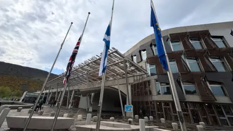 Three flags hanging at halfmast from flagpoles outside the Scottish Parliament, which is in the background. 