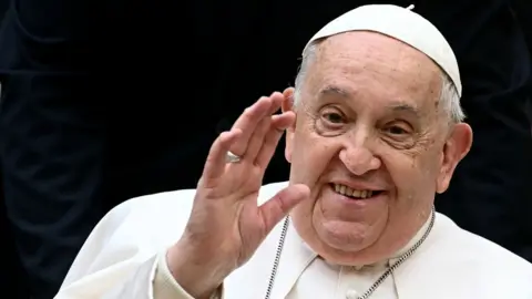 Getty Images The pope, wearing all white, waves with his right hand. There is a black background behind him 