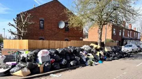 A residential street with a long pile of rubbish stretching along part of it. There are lots of black bin bags as well as boxes and a shopping trolley.