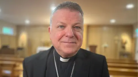 A bishop in a close-up shot in a church. He has short grey hair and is wearing a silver necklace. 