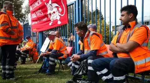 PA Media A group of bin workers, all wearing orange hi-vis jackets, sit on chairs in front of blue railings with a large red protest sign attached to them.