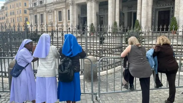 A group of nuns on the left and two women on the right standing behind railings in the Vatican.