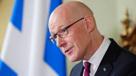 PA Media A bald man with glasses in a close-up shot. He is wearing a dark suit, white shirt and purple tie, with a saltire badge on his jacket. There is a saltire hung in the background.