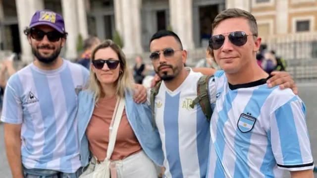 A group of people, three men in blue and white Argentinian football shirts and a woman in a blue shirt and orange tshirt in the Vatican.