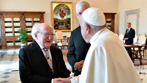 Getty Images The pope is shaking Michael D Higgins hand. Another man stands close to them. A man stands in the background. There is a painting depicting a biblical scene in the background. 
