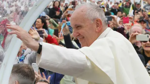 Philip Massey Pope Francis, wearing white formal dresswear, smiles and waves. He has a large crucifix around his neck. There is a crowd on his right, looking at him. 