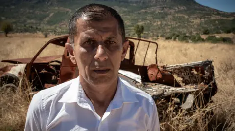 Phil Caller A middle-aged man in a white shirt stands in front of a burned-out vehicle in a field of straw, with mountains in the distance. 