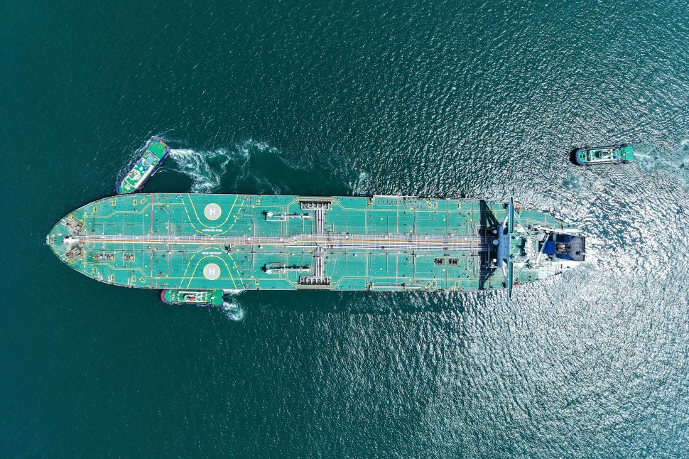 Aerial view of a massive oil tanker being guided by tugboats through deep blue waters, symbolizing the scale and complexity of global energy logistics.