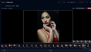 A woman with long, curly hair poses dramatically. She wears a red strappy top and has red lipstick. Her hands are gently framing her face. The background is dark, emphasizing her features.