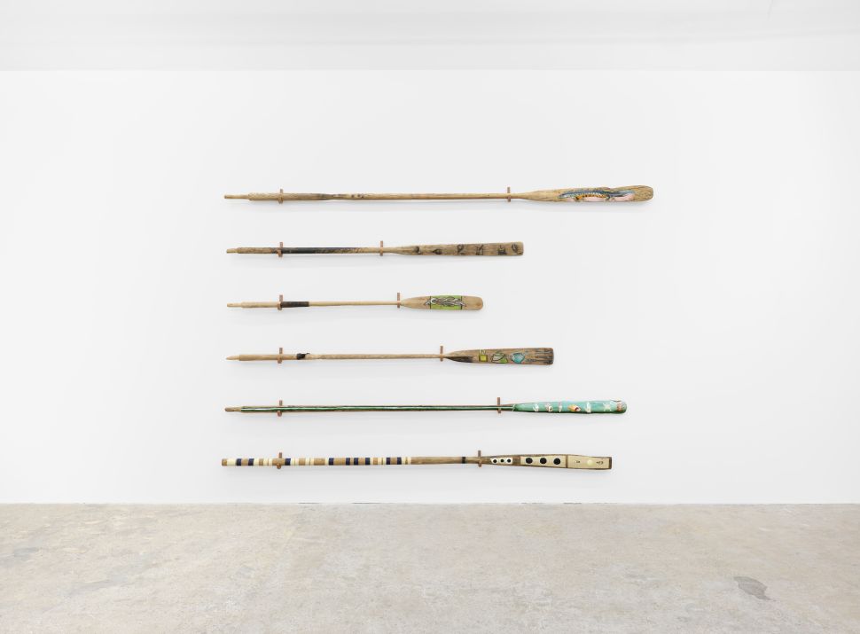 An image of six wooden oars mounted horizontally on a white gallery wall, each one painted or wrapped with different patterns, textures, and materials as part of Lubaina Himid’s Barricades installation at Hollybush Gardens in London.