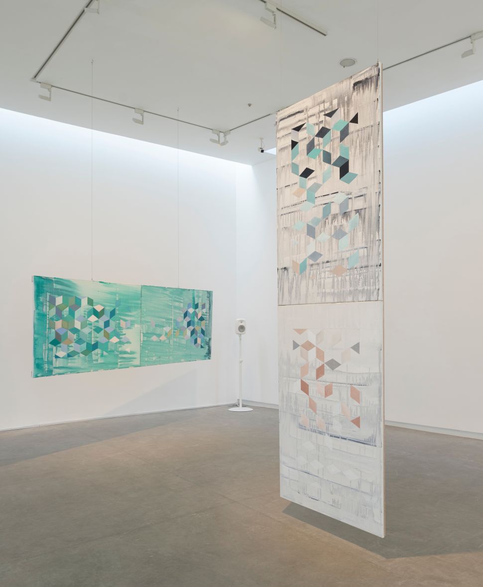 An image of three large abstract geometric paintings—two teal and one in grayscale—suspended from the ceiling in a brightly lit gallery space, part of Zanzibar (1998–2023) by Lubaina Himid and Magda Stawarska, featuring Stawarska’s audio installation component in the background.