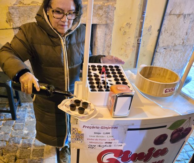 a woman in a warm coat pouring liquor into little chocolate cups. her stand reads 'Ginja' an alternate spelling of ginjinha