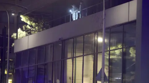 Reuters Two people wearing white forensics suits are seen on the roof of a building, they are shining a torch and looking down.