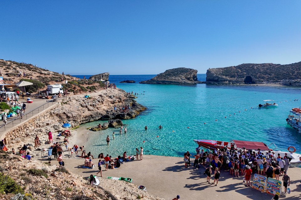 Aerial view of Blue Lagoon, Camino, Malta, showing many people swimming and relaxing on the beach and rocky coastline.