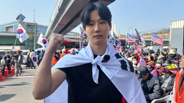 Kim Dong-hyeon, wearing the Korean national flag on his back, holds a fist up in front of the camera