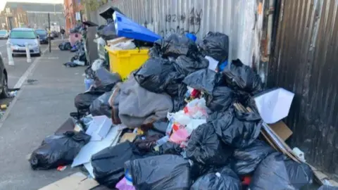 A pile of rubbish, including black bin bags and what looks like a grey carpet or quilt. Large wheelie bins nearby are bulging open from being so full.