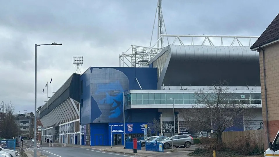 Portman Road football stadium, the picture is taken of the corner of the stadium, a large image of Sir Bobby Robson is on the stadium wall, above the entrance to the club's shop. Floodlights are seen above the top of the stadium. Cars are parked close by in a car park.