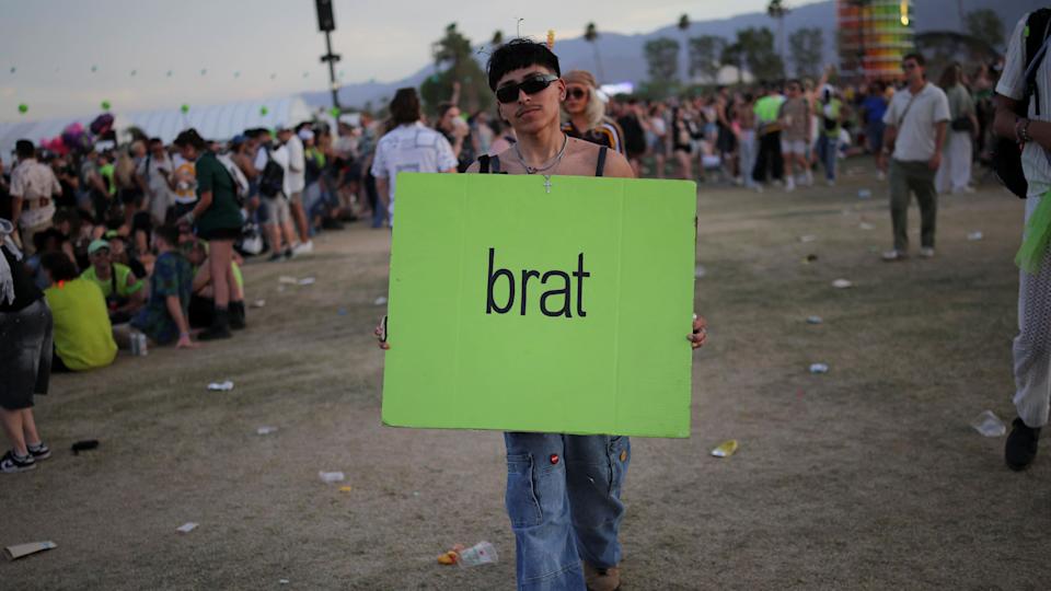 A fan wearing sunglasses and holding a large piece of cardboard, painted green with the words 'Brat' written on it.
