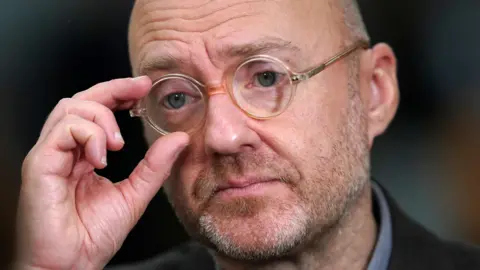 PA Media A bald man adjusts his glasses with his right thumb and index finger in a close-up shot. He has as short stubbly beard and is looking to the left of the camera, with a dark background out of focus behind him. 