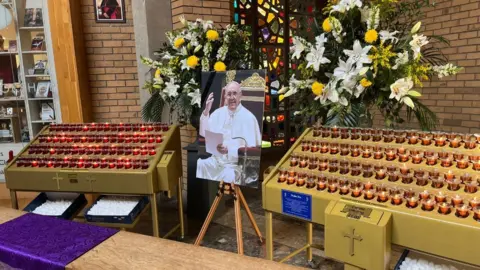 A picture of the pope sits on a stand between rows candles in gold cases, with Christian crosses on the front. There are flowers and a stained-glass window behind