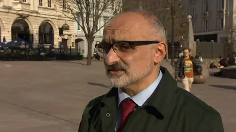 Onay Kasab has very short grey and black hair and is wearing black glasses. He has grey facial hair and is wearing a dark green coat with a light blue shirt and red tie.