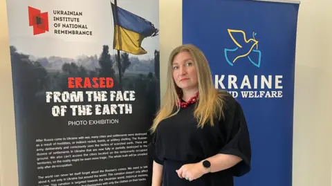 BBC Antonina Grebeniuk stands in front of two banners at the Erased from the Face of the Earth exhibition at the M Shed in Bristol.
