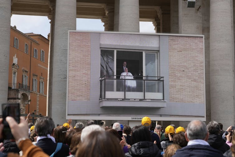 A crowd of people, seen from the back, look up toward a large screen that shows a man in white sitting on a small balcony.