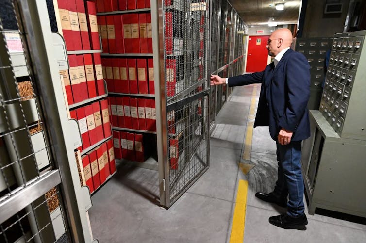 A man in a blue suit swings open a door to storage shelves full of large red books.