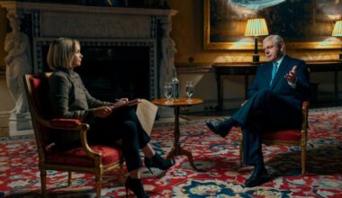 A short-haired woman in a grey top, trousers and heeled boots sits opposite a middle-aged man in a navy suit and tie, as if in an interview. They both sit on opulent upholstered chairs in front of a large fireplace.