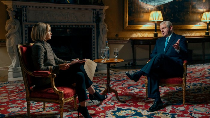 A short-haired woman in a grey top, trousers and heeled boots sits opposite a middle-aged man in a navy suit and tie, as if in an interview. They both sit on opulent upholstered chairs in front of a large fireplace.