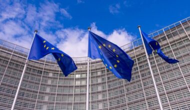 Three European Union flags wave in front of a modern glass building under a blue sky