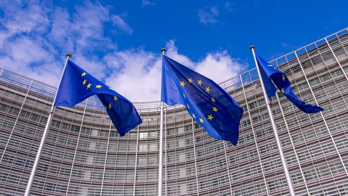 Three European Union flags wave in front of a modern glass building under a blue sky