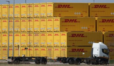 A white truck is parked in front of a large stack of yellow DHL containers at a logistics center. The containers are labeled with the DHL logo and the slogan &quot;Excellence. Simply Delivered.&quot;