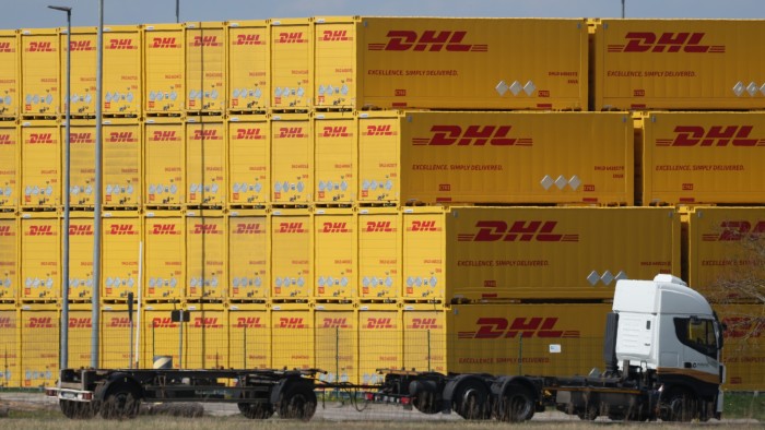 A white truck is parked in front of a large stack of yellow DHL containers at a logistics center. The containers are labeled with the DHL logo and the slogan &quot;Excellence. Simply Delivered.&quot;