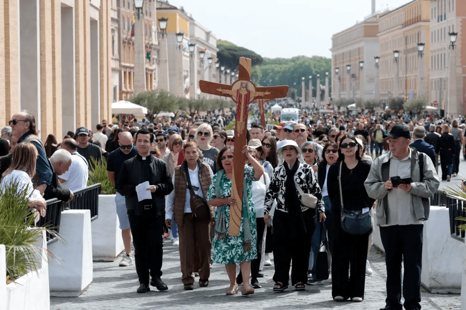 A large crowd of people walking down a street, carrying a large wooden cross.