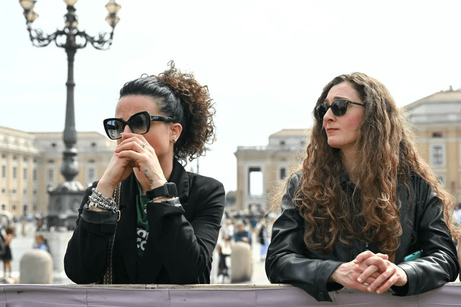 Two women wearing sunglasses stand behind a barrier, one appearing upset.
