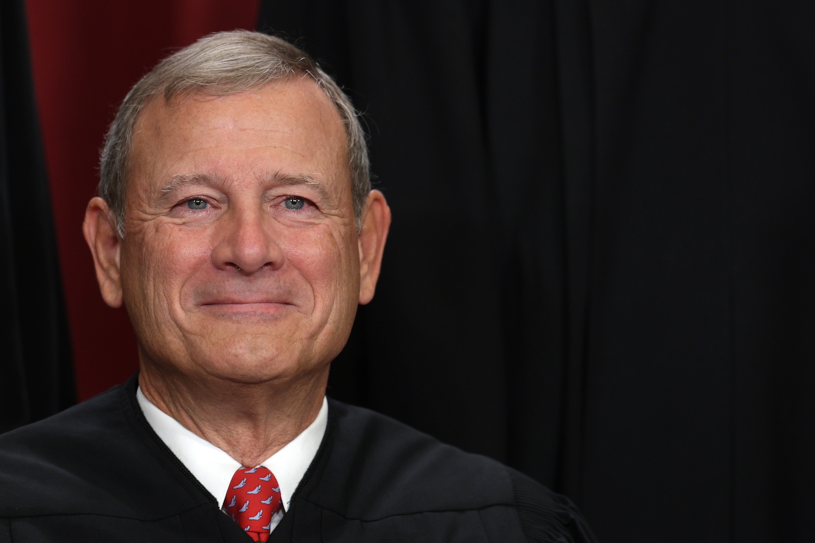 A close-up of a white man's face. He is smiling tightly, has gray hair, and is wearing a red tie