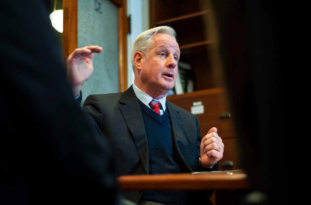 Man in a suit gesturing while speaking, seated at a table in an office setting.