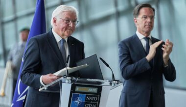 Steinmeier steht lächelnd an einem Rednerpult. Daneben Rutte, der applaudiert. Im Hintergrund sieht man unscharf eine Glasfassade und einen Mann