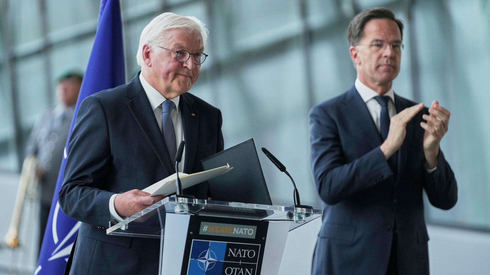 Steinmeier steht lächelnd an einem Rednerpult. Daneben Rutte, der applaudiert. Im Hintergrund sieht man unscharf eine Glasfassade und einen Mann