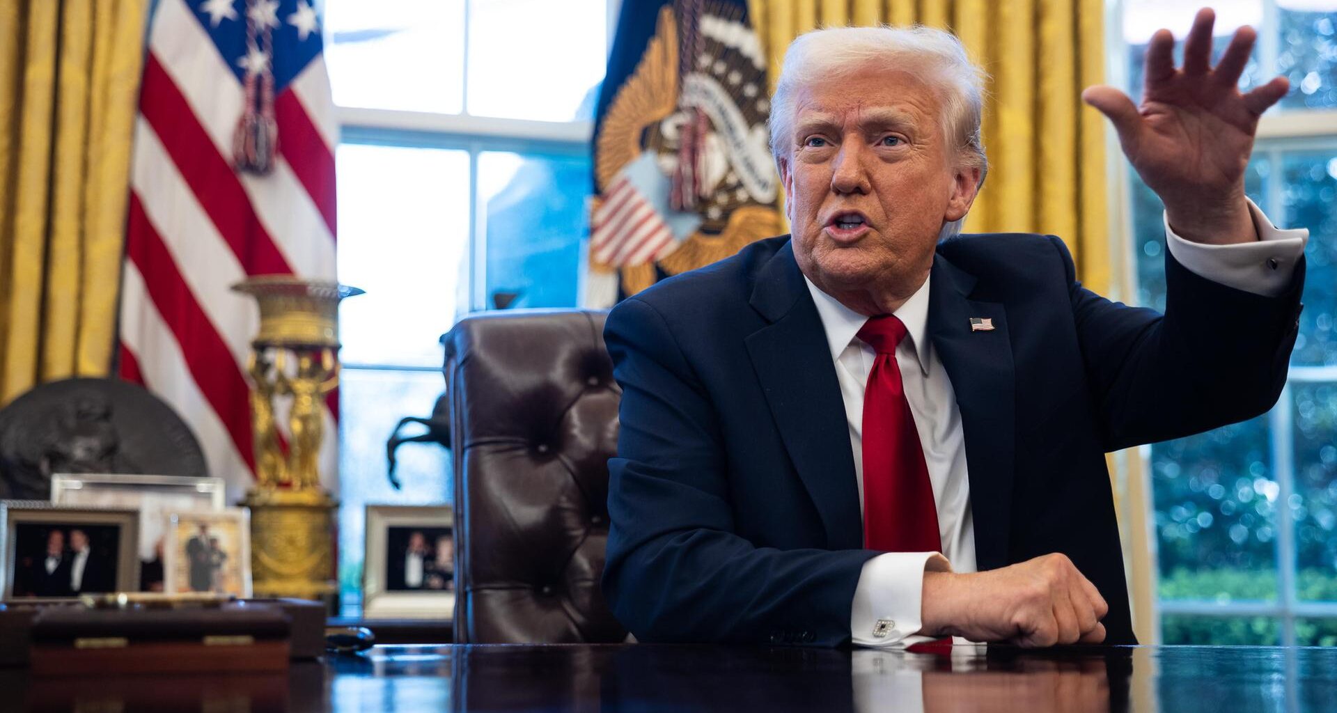 US-Präsident Donald Trump im Anzug mit roter Krawatte im Weißen Haus am Schreibtisch. Hinter ihm eine US-Flagge. Er gestikuliert gut gelaunt mit einer Hand.