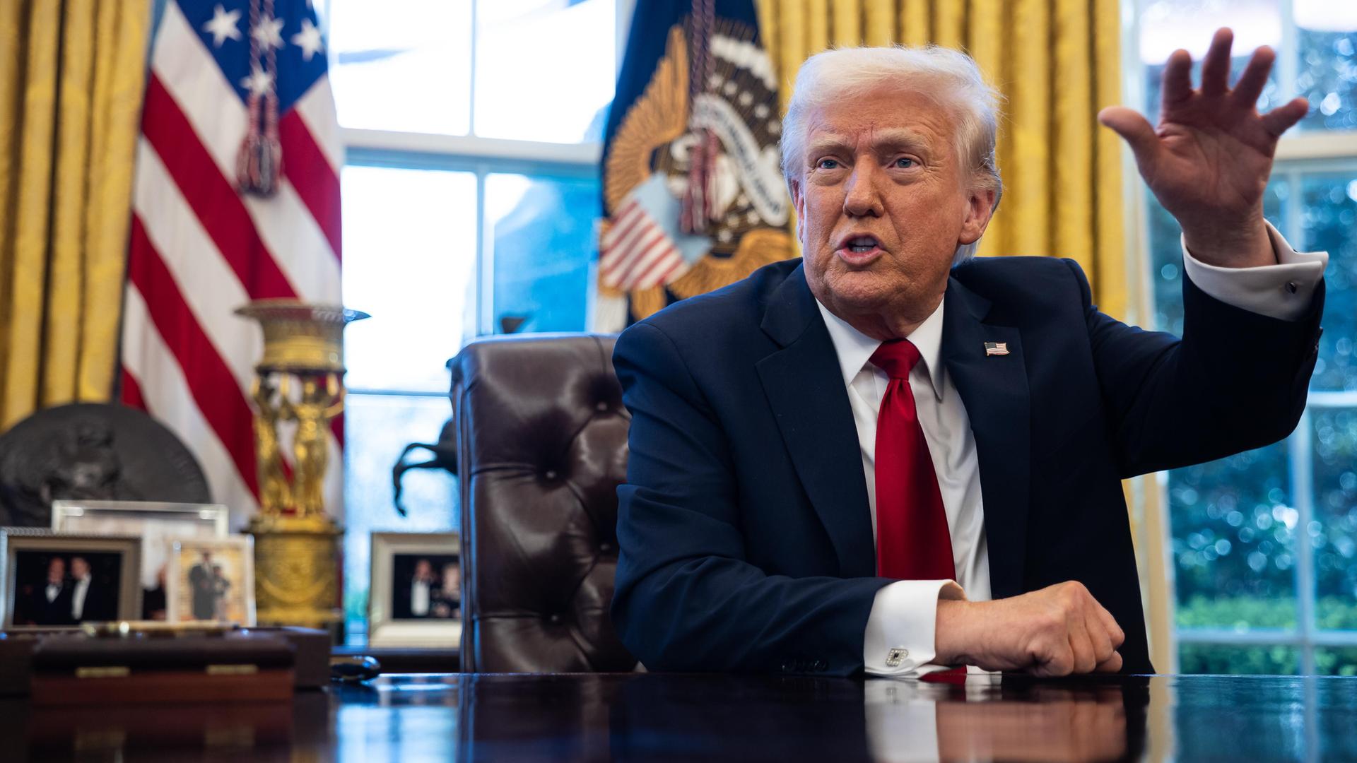 US-Präsident Donald Trump im Anzug mit roter Krawatte im Weißen Haus am Schreibtisch. Hinter ihm eine US-Flagge. Er gestikuliert gut gelaunt mit einer Hand. US-Präsident Donald Trump im Anzug mit roter Krawatte im Weißen Haus am Schreibtisch. Hinter ihm eine US-Flagge. Er gestikuliert gut gelaunt mit einer Hand.