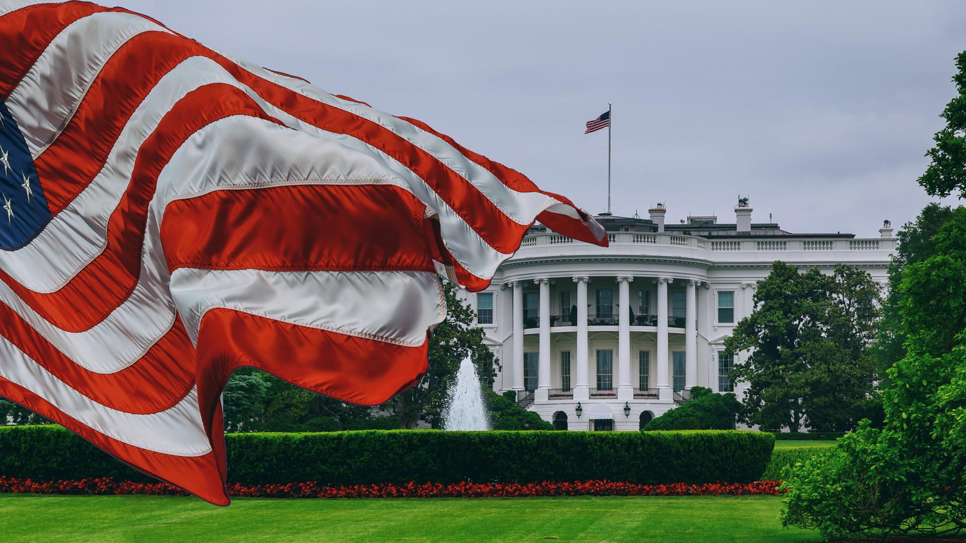Zu sehen ist das Weiße Haus, im Vordergund weht eine US-Flagge. Zu sehen ist das Weiße Haus, im Vordergund weht eine US-Flagge.
