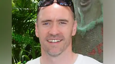 BBC Paul Doyle has short dark greying hair with sunglasses on his head and is wearing a white T-shirt.